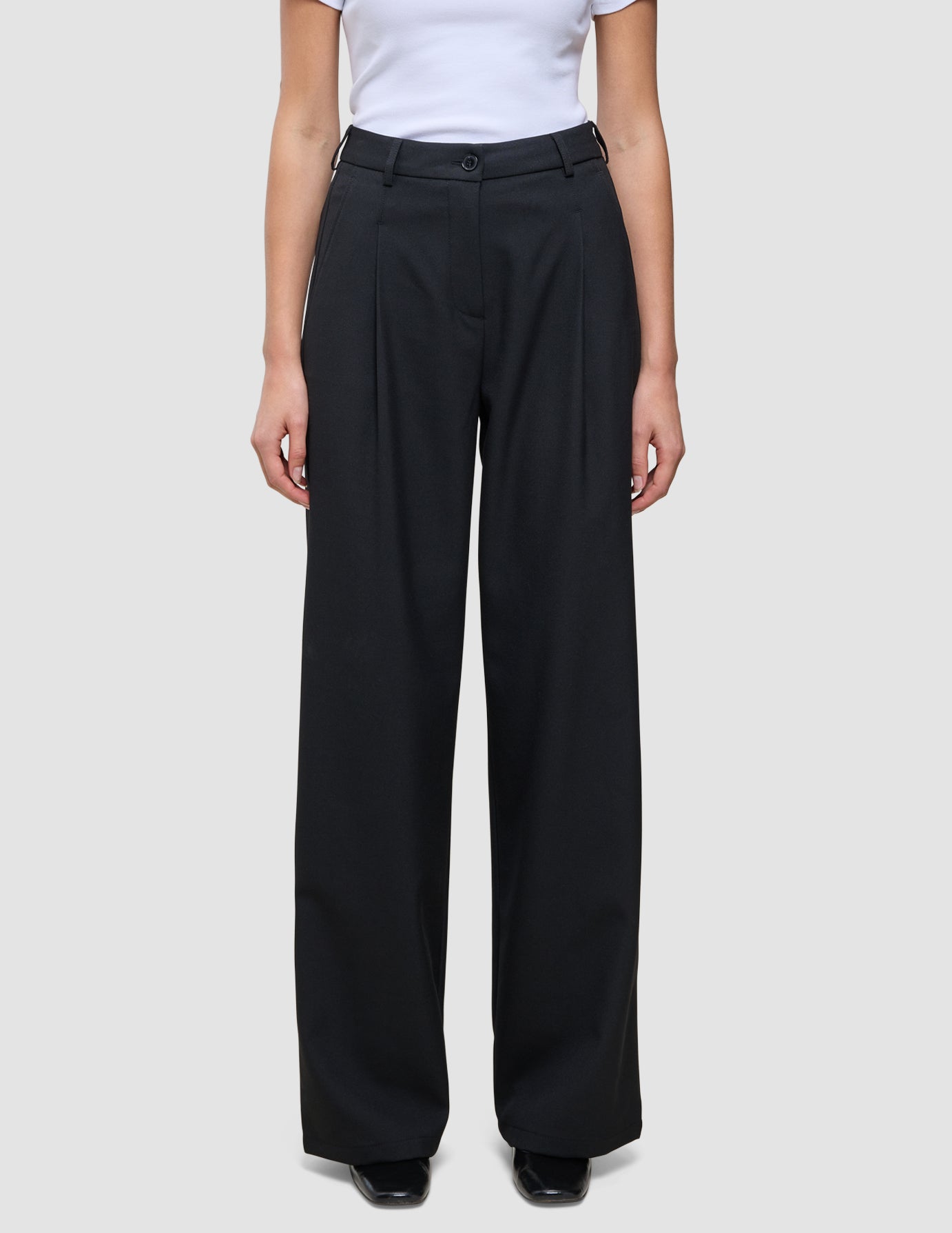 En person som har på seg de svarte Serene Pants Wide Black-buksene med plissering foran og en hvit T-skjorte i skandinavisk stil.
